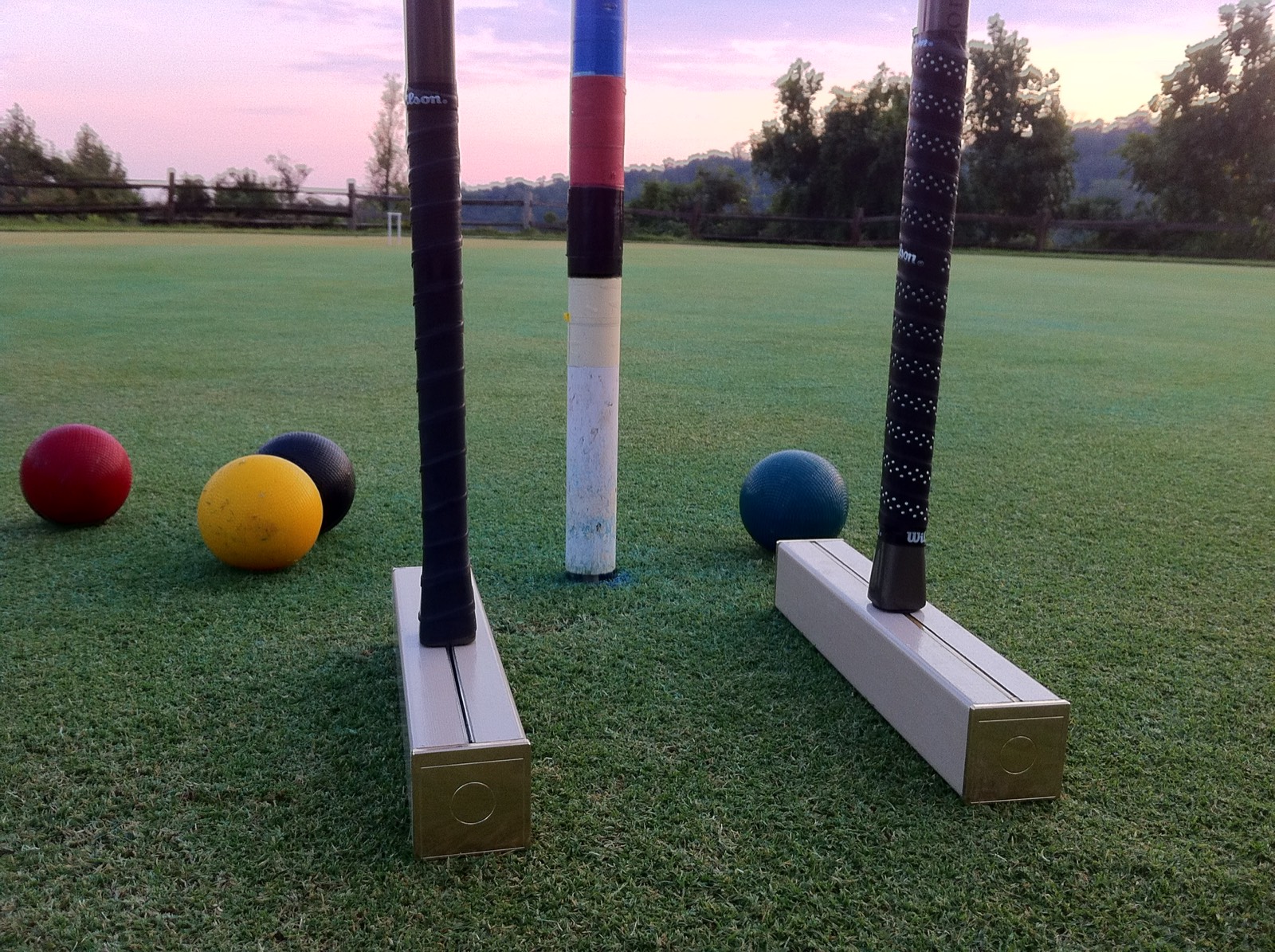 Two mallets stand on end beside the striped peg, with red, yellow, black and blue balls on the lawn under a pink sky.