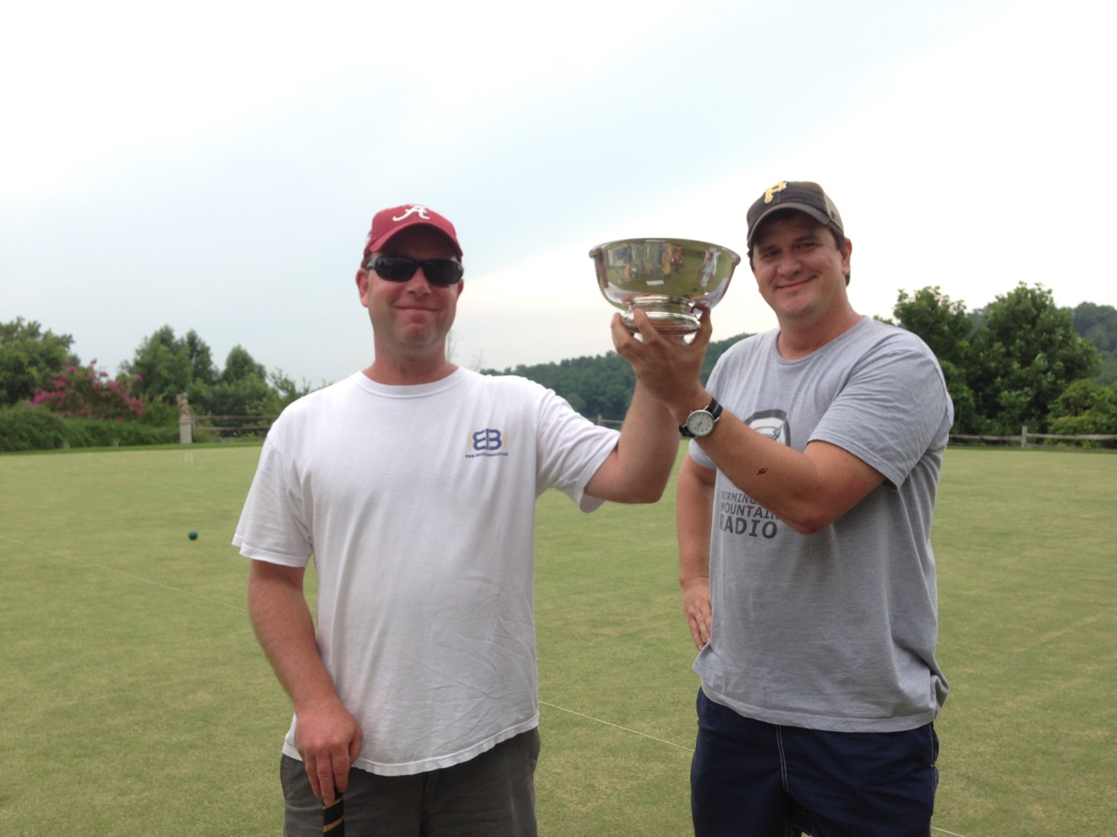 Two club members raise a silver trophy bowl together on a croquet lawn, with a ball and a hoop behind them.