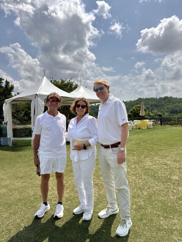 Three people dressed in white smile on the lawn, with white event tents and yellow-draped tables behind them.