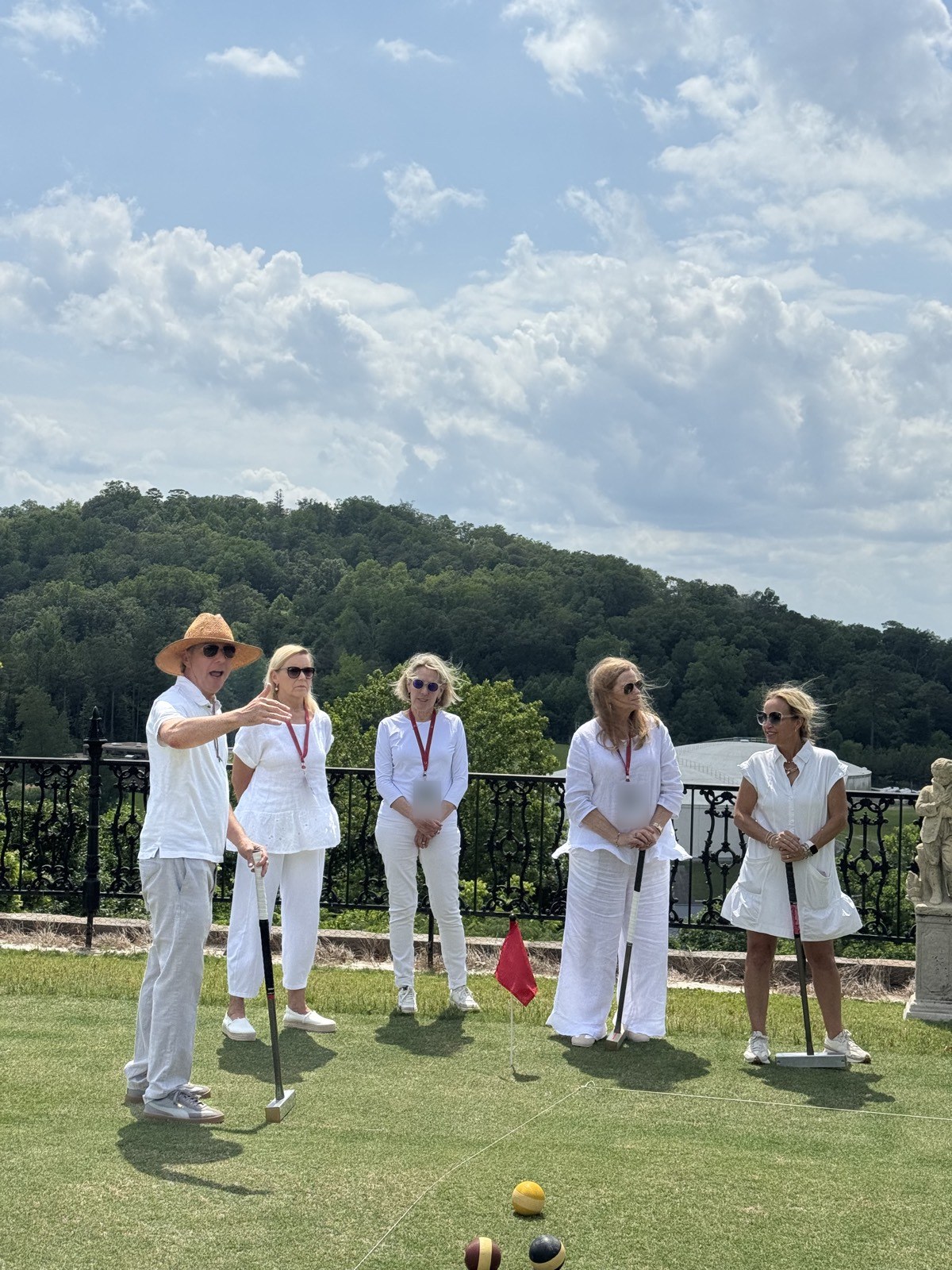 A player in a straw hat explains the game to four women in white on a lawn, two of them holding mallets.