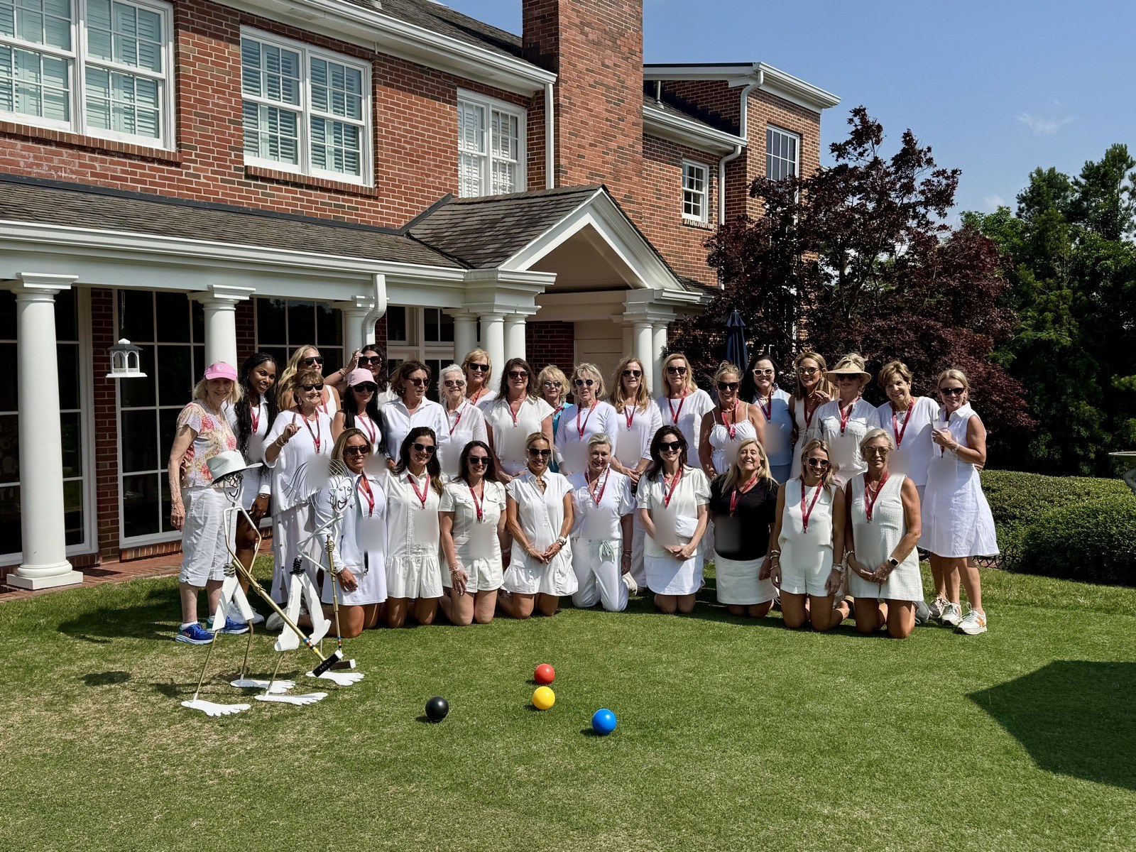 A large group of guests in white poses on the Shook Hill lawn behind croquet balls and a metal sculpture of two players.