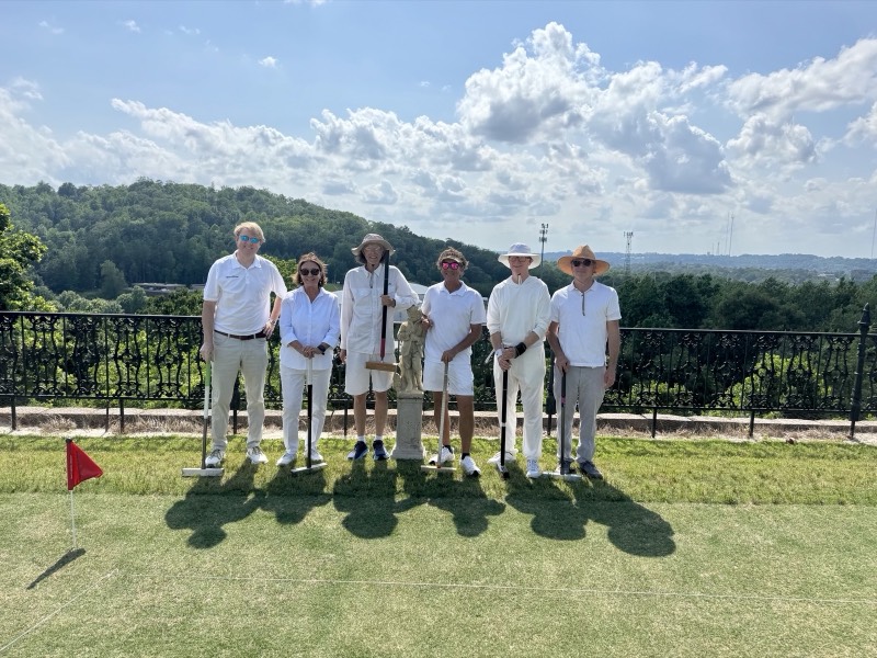 Six players in white stand with their mallets on a lawn beside an iron railing, with wooded hills behind them.