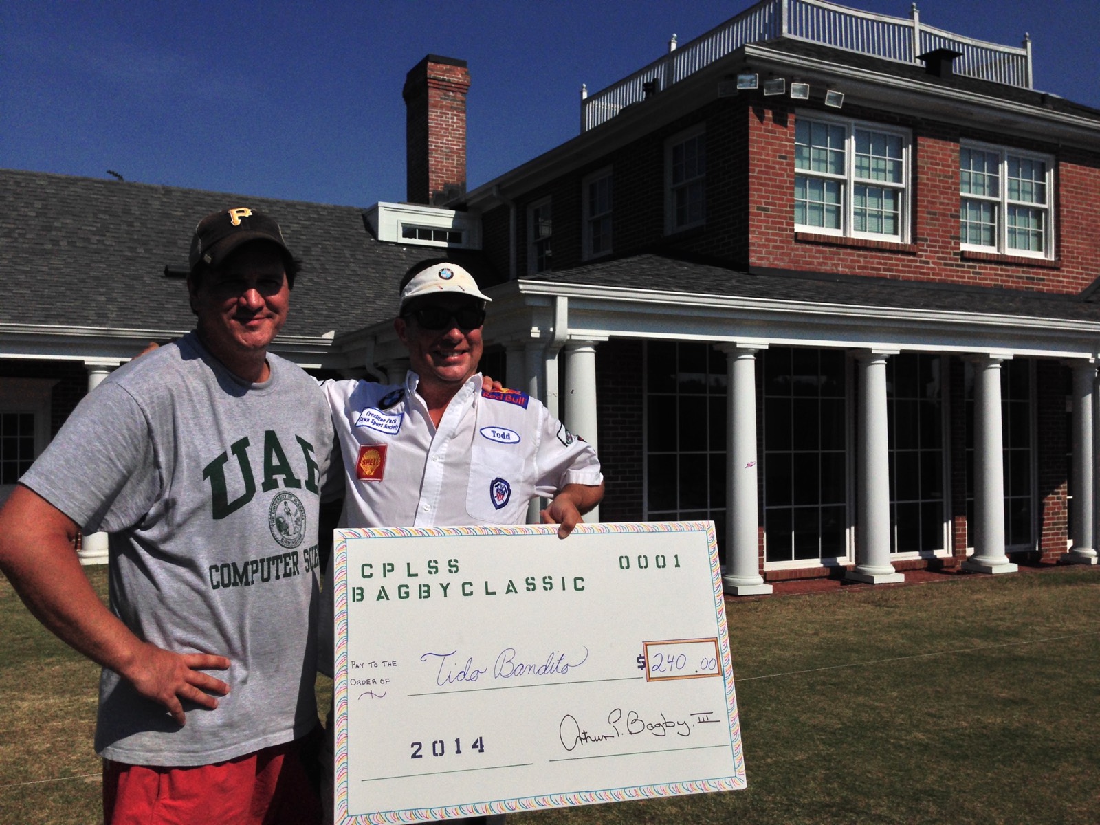 Two players pose with an oversized CPLSS Bagby Classic prize check on the lawn in front of a large brick house.