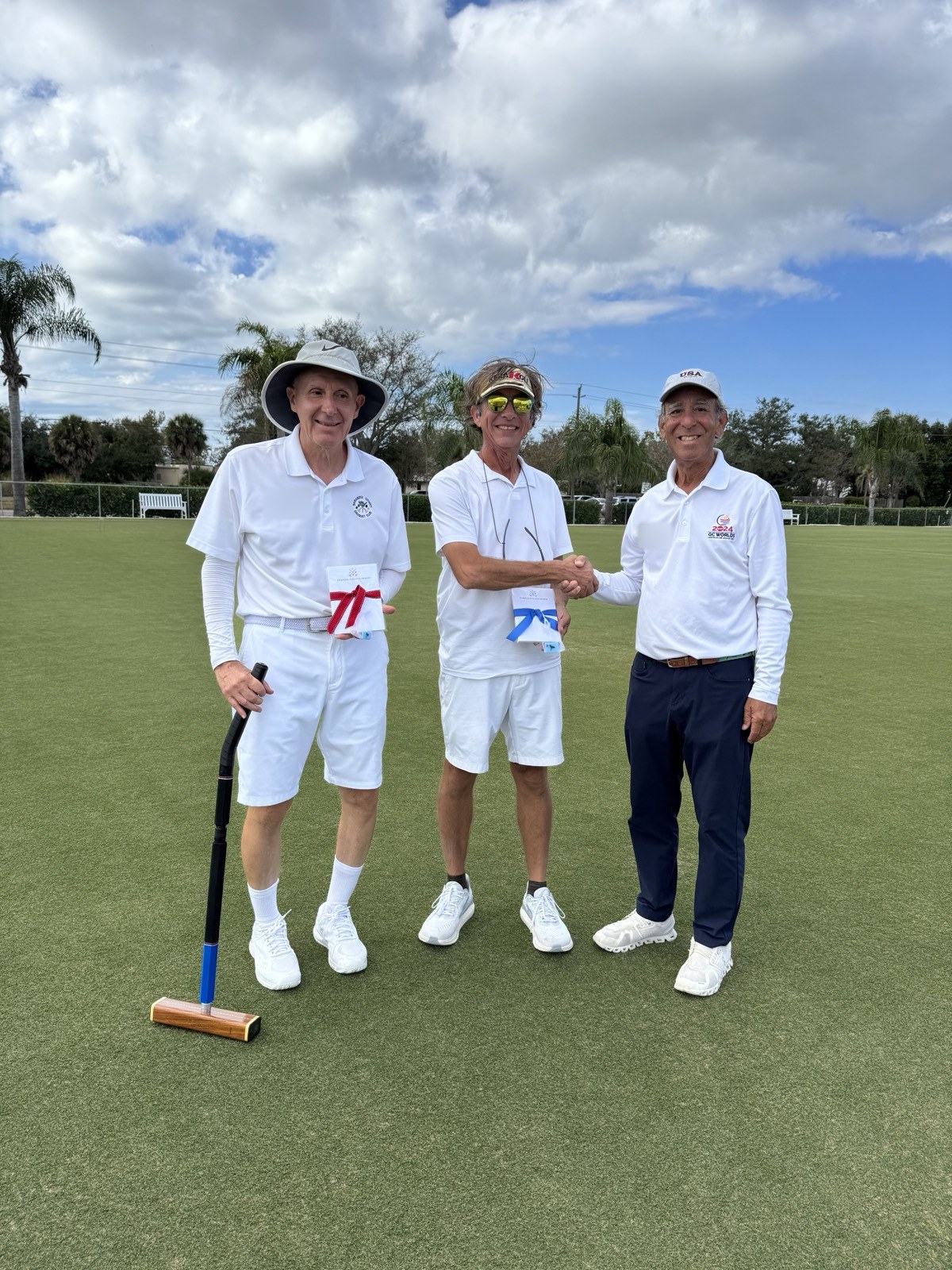 Two players holding small ribbon-tied gifts stand on a croquet lawn as one shakes hands with a man in a USA cap.