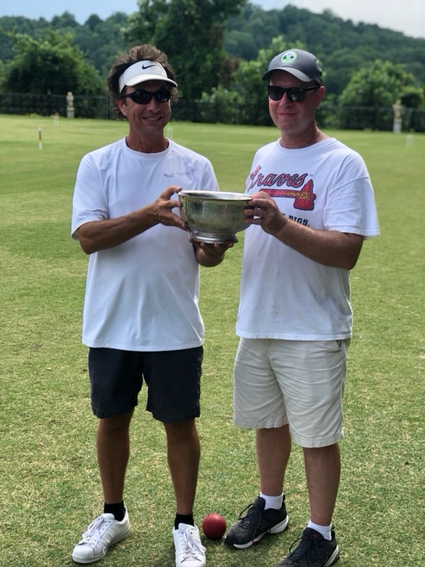 Two players smile as they hold a silver trophy bowl between them on a croquet lawn, with trees and a fence behind.