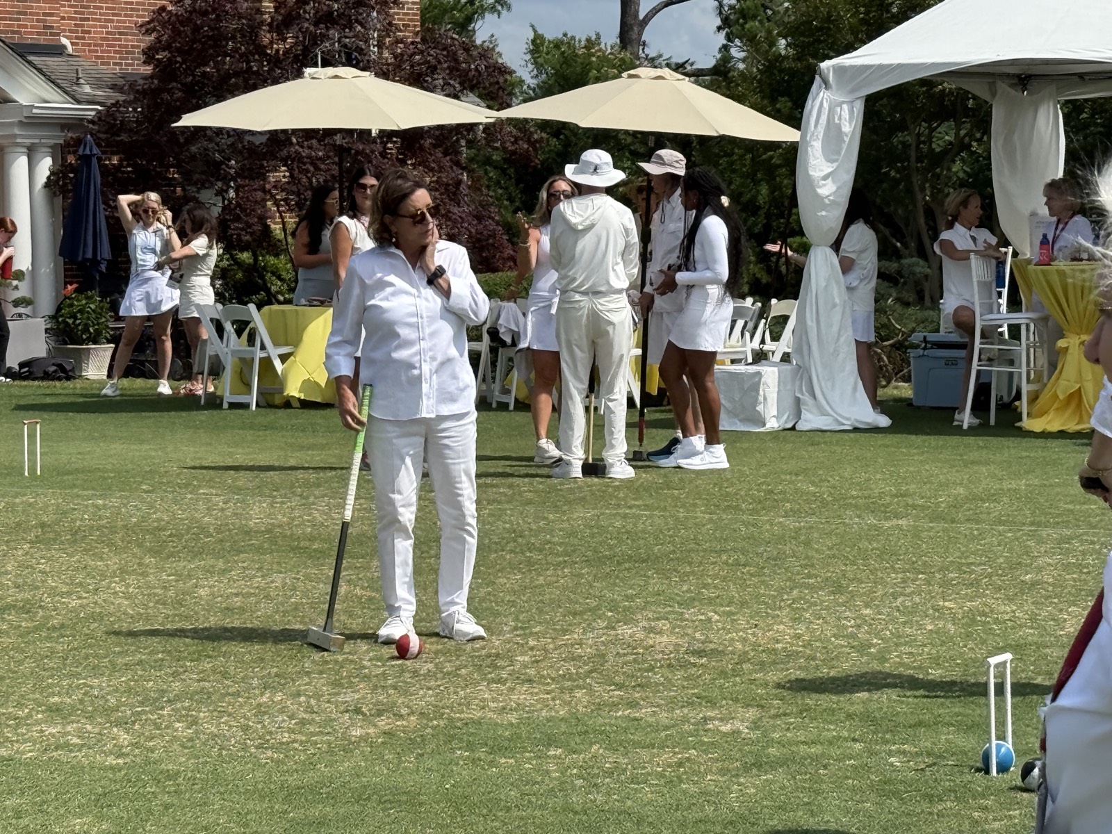 A player in white stands with her mallet by a red ball as guests gather under umbrellas and a white tent.