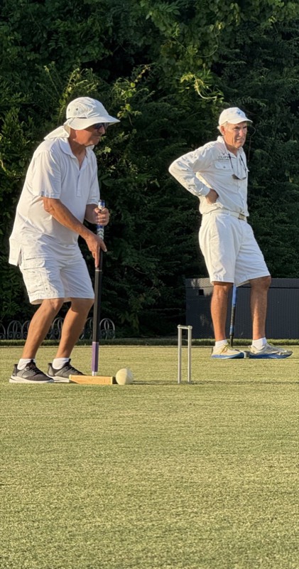 A player in white lines up his mallet behind a ball near a hoop while another player watches, one hand on his hip.