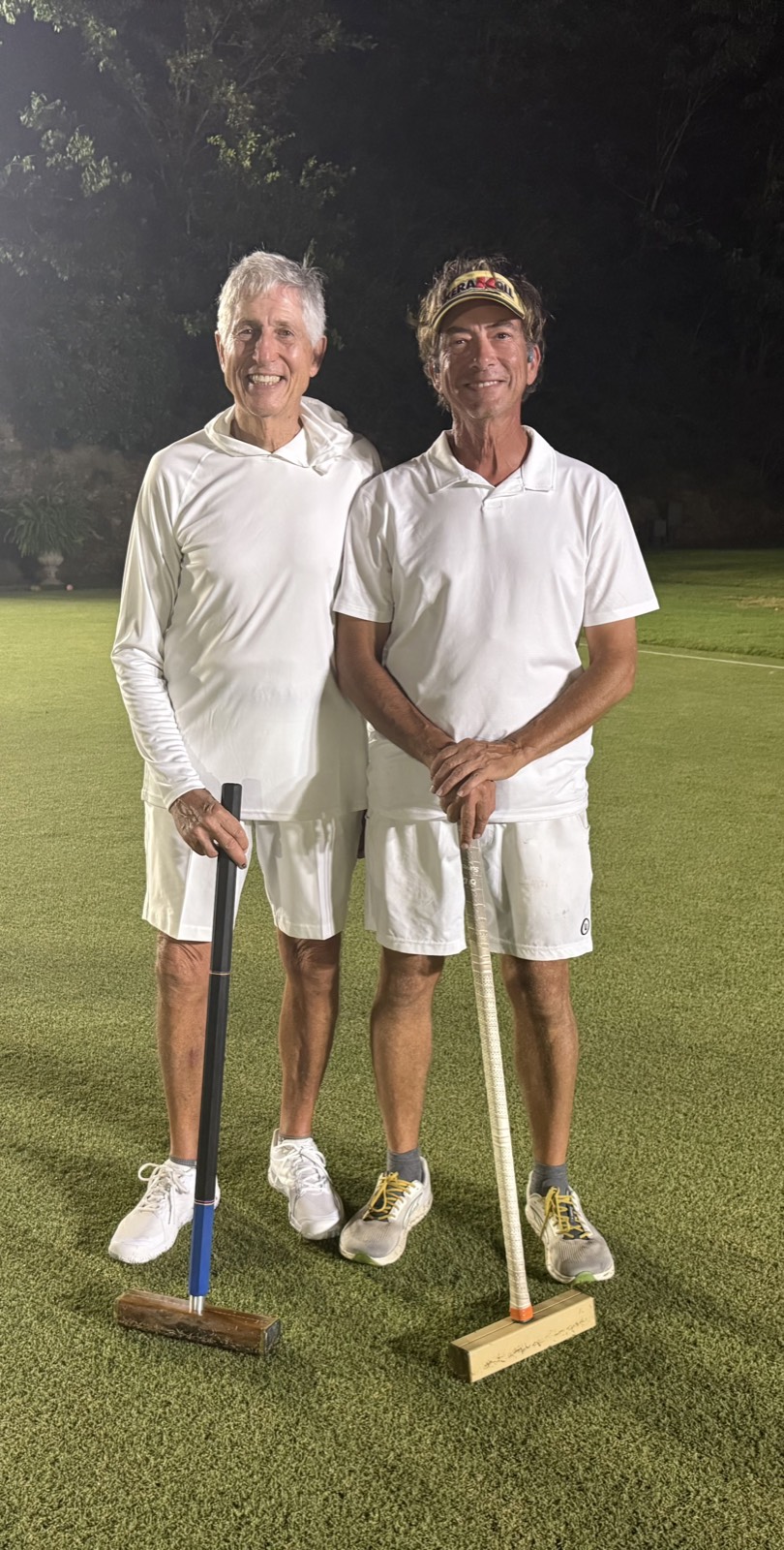 Two players in whites lean on their mallets on the lawn at night, under the lights at the 2025 Golf Croquet Open.