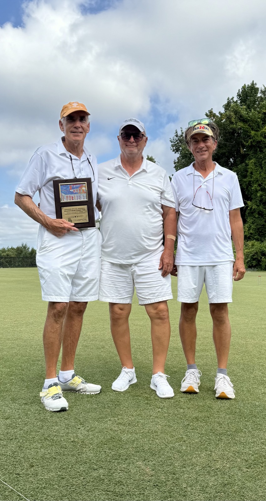 Three players in croquet whites stand together on the lawn, the one on the left holding a plate champion plaque.