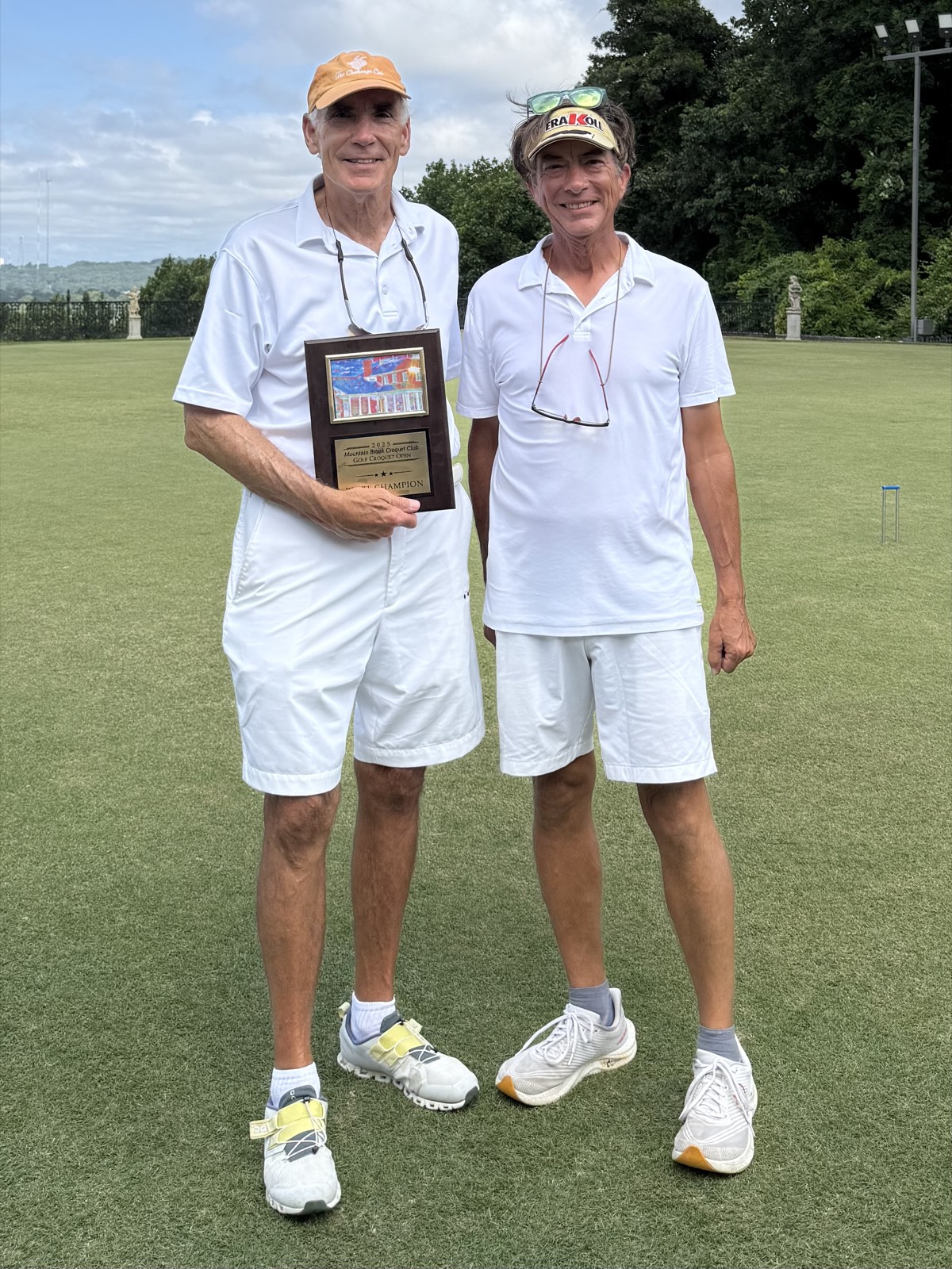 Two players in croquet whites stand on the lawn, one holding a plate champion plaque from the 2025 Golf Croquet Open.