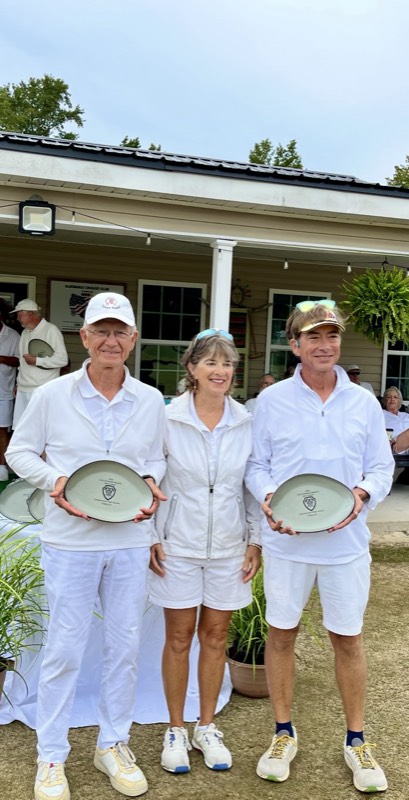 Three players in white pose in front of a clubhouse porch, two holding award plates, as spectators look on behind.
