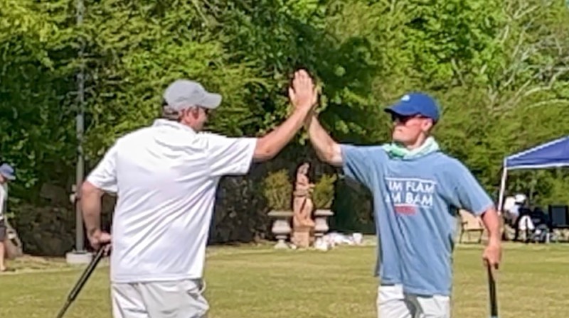 Two players holding mallets trade a high five on a sunny croquet lawn, with a garden statue and trees behind them.