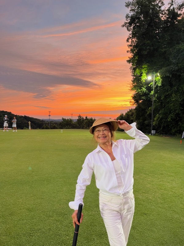 A player in white tips her straw hat and leans on her mallet on a croquet lawn under an orange sunset sky.