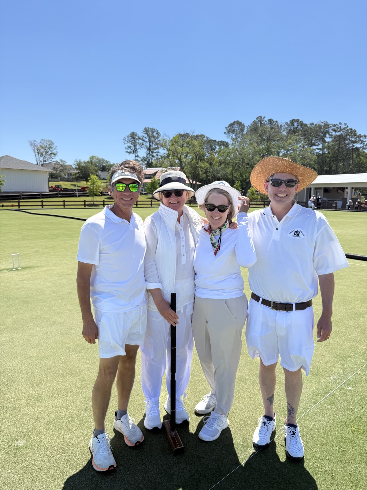 Four players, mostly in white, pose together on a croquet lawn, one of them holding a mallet.