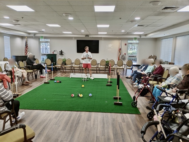 A club member holding notes speaks at the end of an indoor carpet court with mallets, balls and a hoop as residents watch.